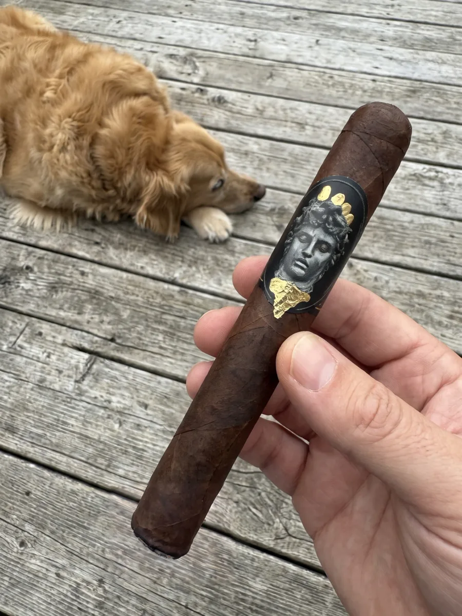 Norm’s hand holding the ALEC & BRADLEY GATEKEEPER TORO 6×52 on the porch, its silver Medusa band in view, with Dallas the golden retriever resting on the deck behind