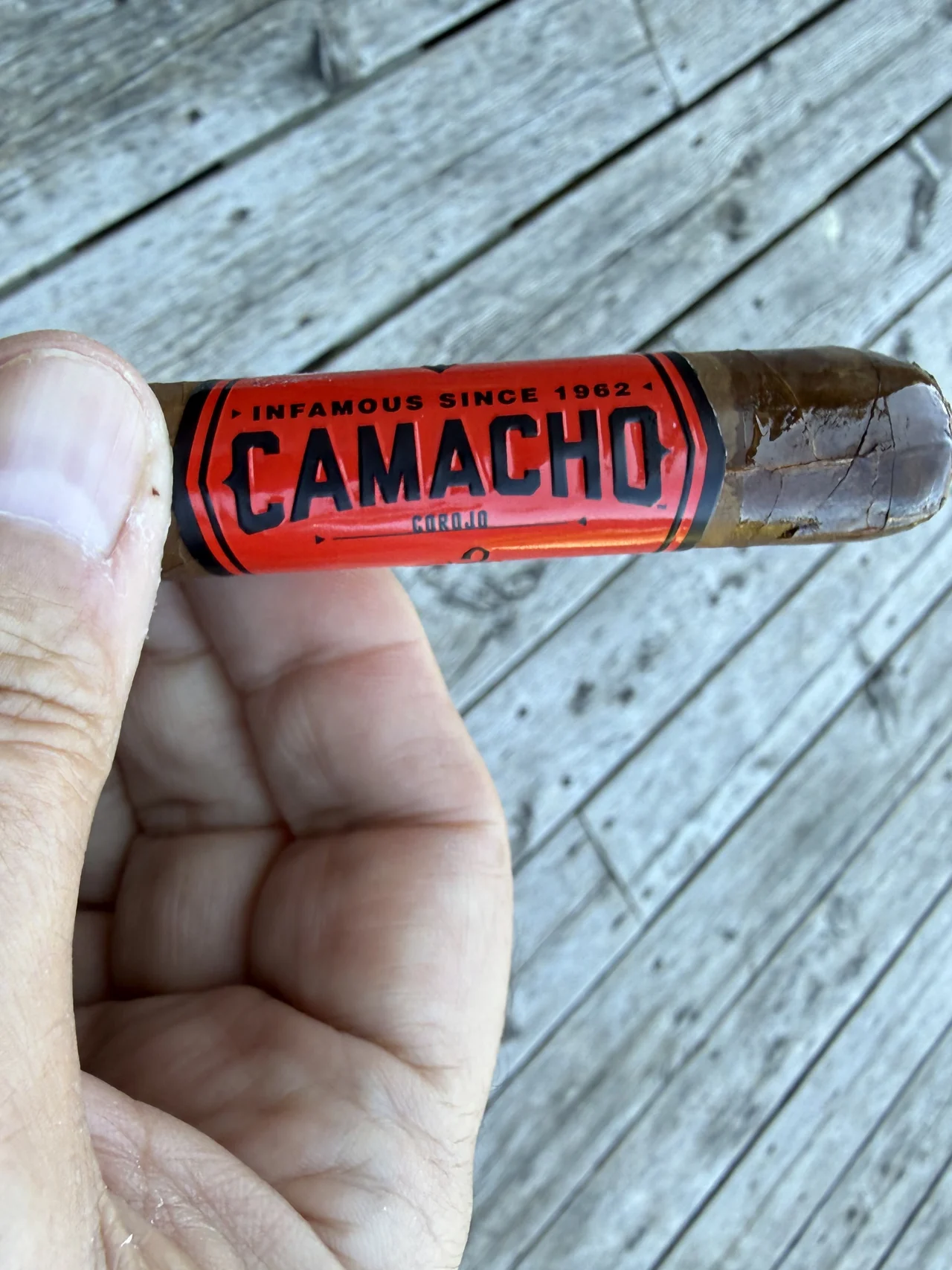CAMACHO COROJO ROBUSTO 5×50 in hand over the grey deck boards