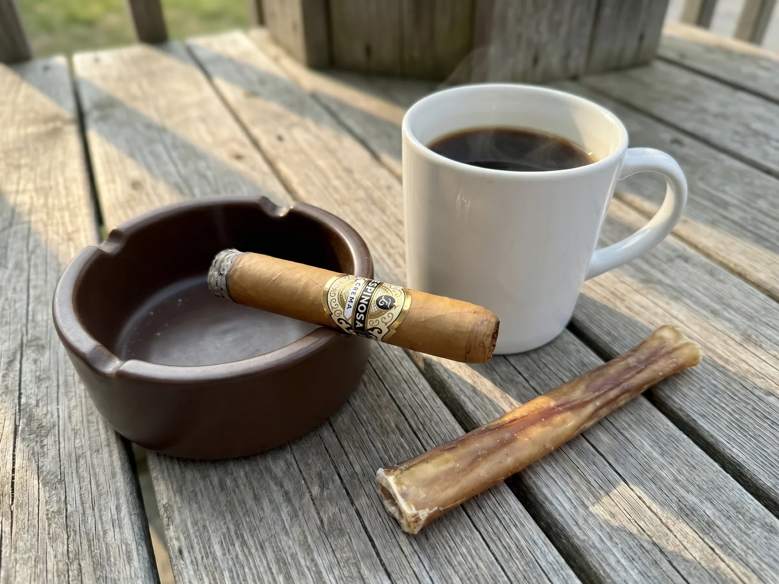 The Espinosa Crema resting on an ashtray beside a cup of black coffee and a bully stick on the porch boards