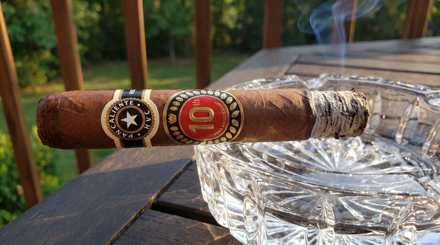 Pan Caliente de HVC 10th Anniversary cigar resting in a large crystal ashtray, grey ash formed at the burn line, black star band and red 10th Year Anniversary band facing the camera