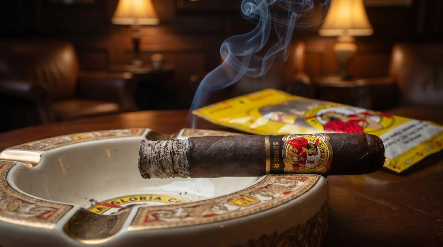 A La Gloria Cubana Serie R resting in a branded La Gloria ashtray with a thin curl of smoke, the yellow fresh pack pouch soft in the background of a warm lounge