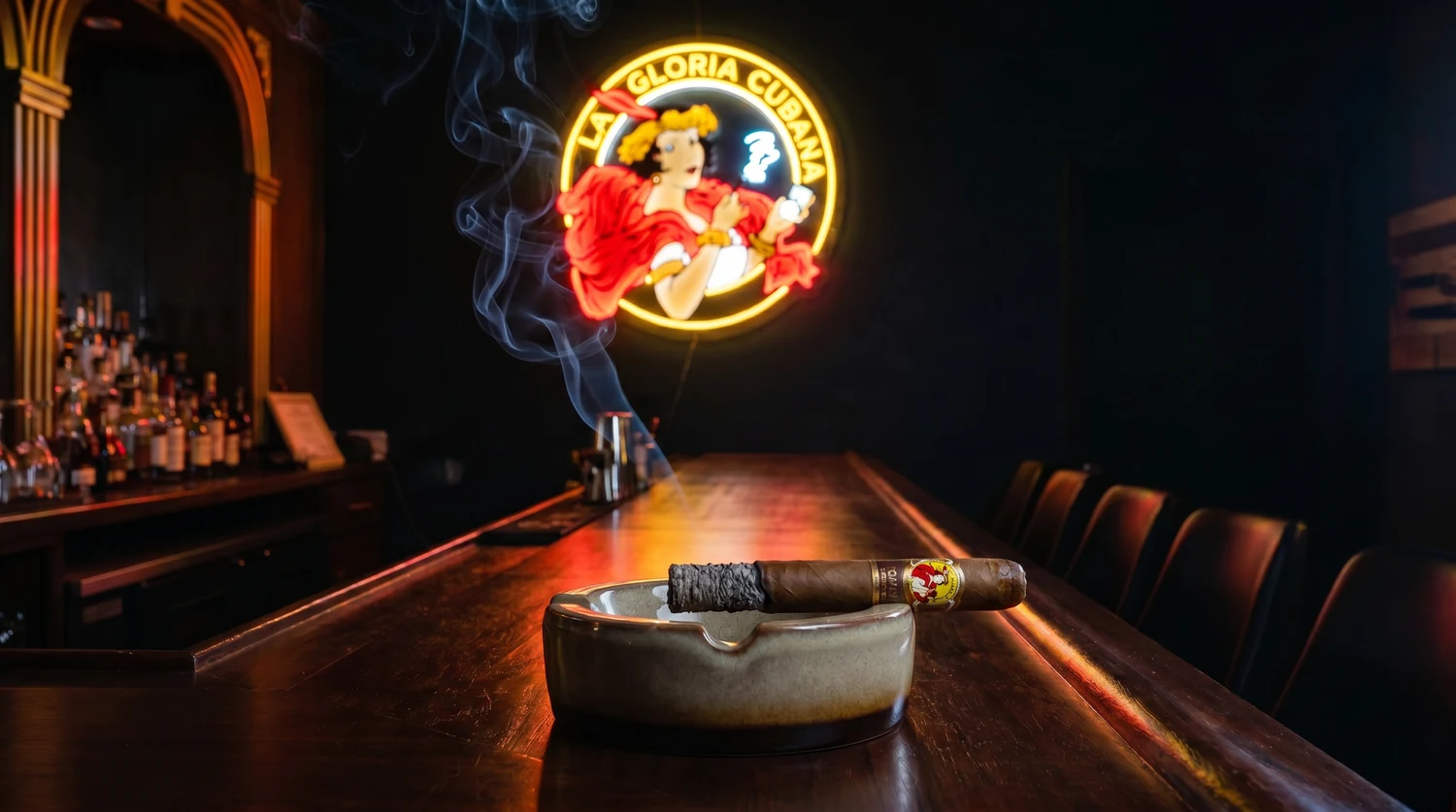 A lit La Gloria Cubana cigar resting in a large ashtray in the foreground of a dark cigar bar, a glowing neon La Gloria Cubana lady sign on the wall behind