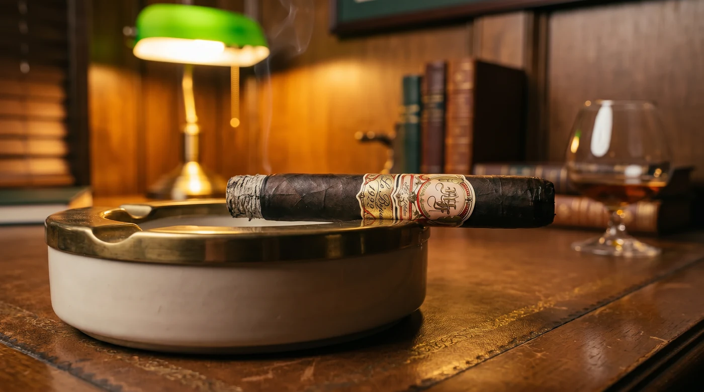 My Father Le Bijou 1922 100 Años resting in a large cigar ashtray on a leather-topped study desk under a banker's lamp, a pour nearby, smoke rising from the foot