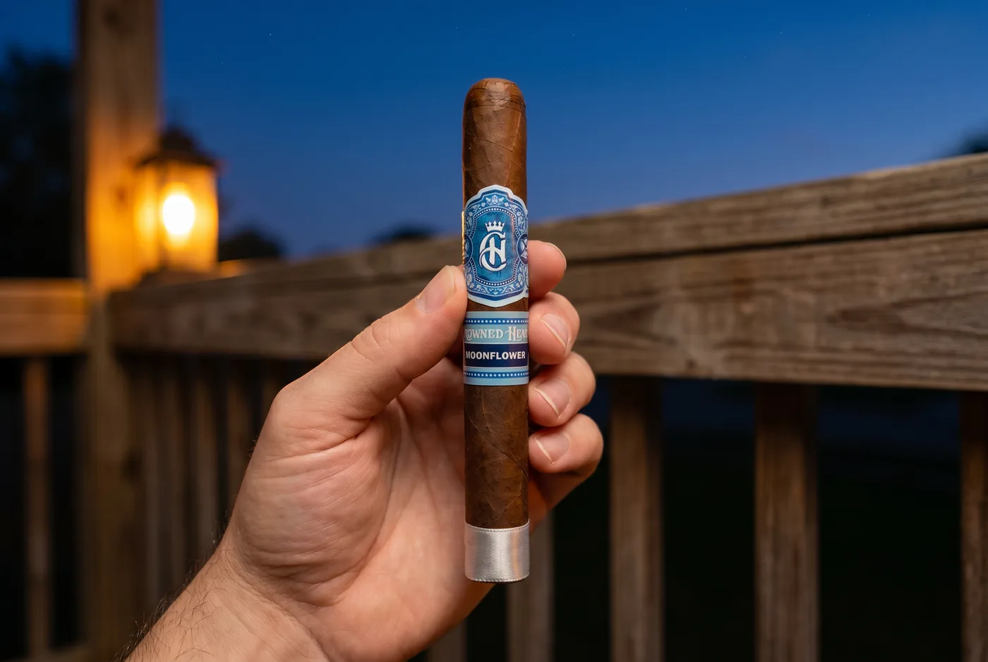 Hand holding a Crowned Heads Moonflower Corona Gorda on a deck at dusk, deep blue band facing out