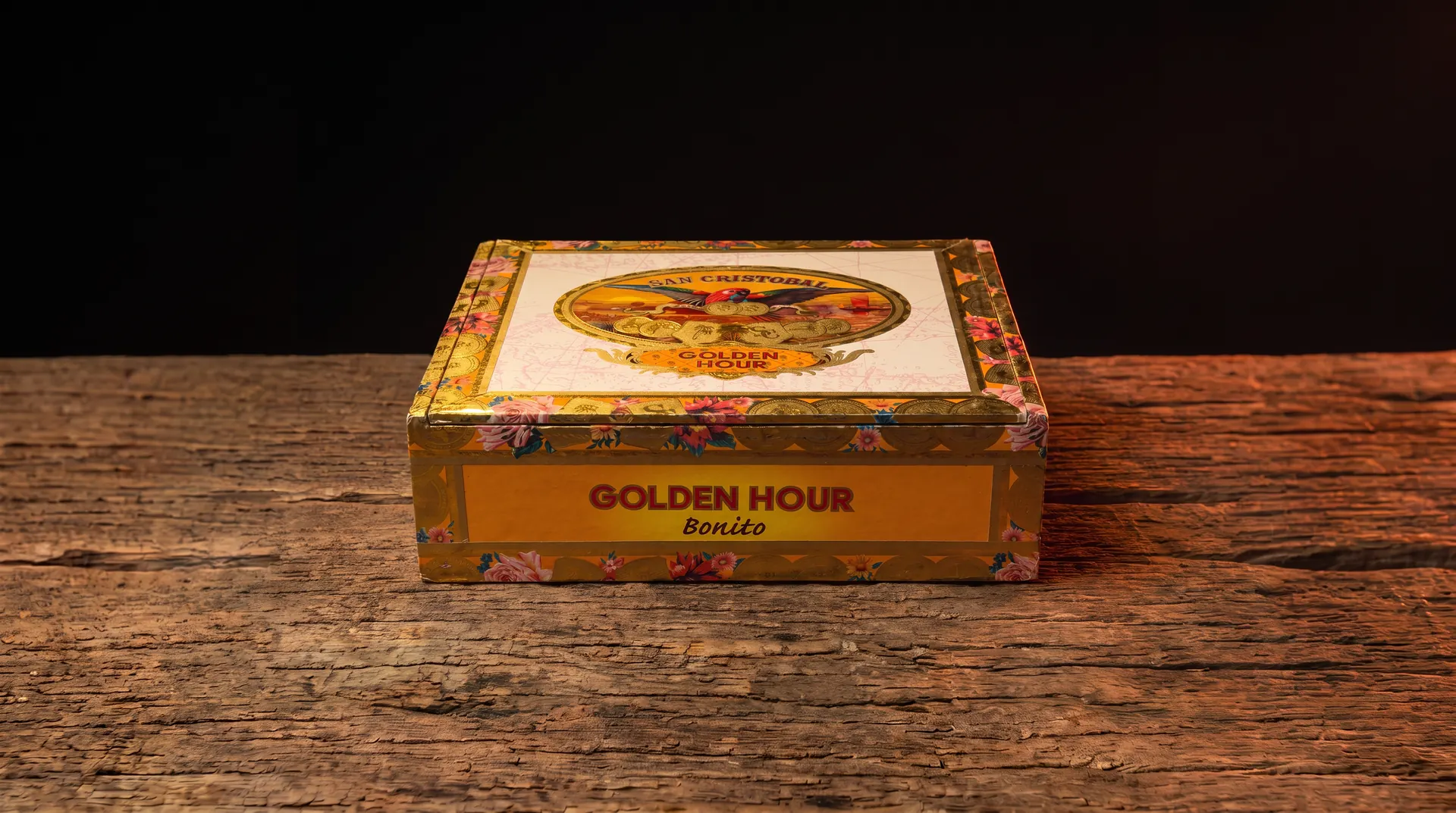 San Cristobal Golden Hour Bonito cigar box, gold and orange with a macaw and coin crest on the lid, resting on a dark wood table