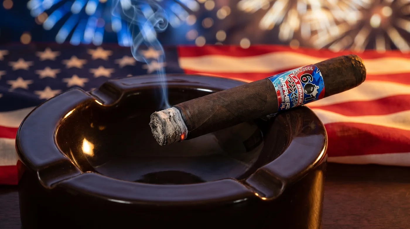 A lit 601 La Bomba Warhead Independence Day resting in a large cigar ashtray, American flag and fireworks behind, smoke rising from the foot