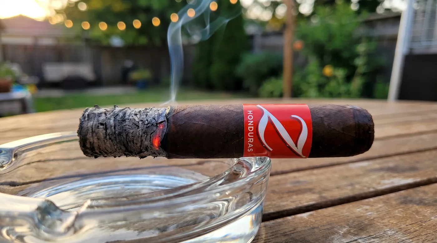 A lit Zino Honduras robusto with a long deep ash on a glass ashtray, smoke rising from the burn line, backyard at golden hour